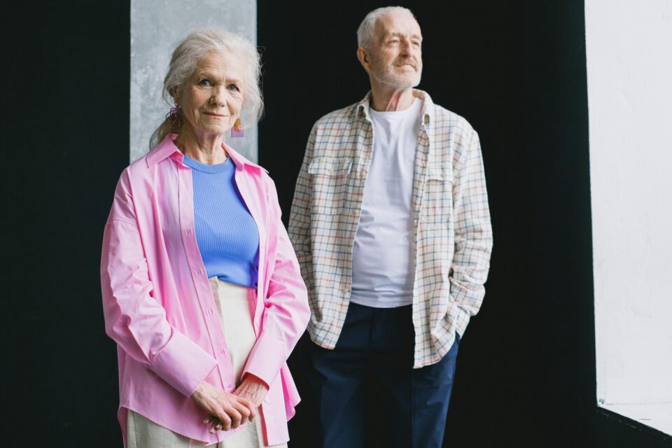 Smiling elderly couple standing indoors wearing colorful casual clothing, exuding warmth and charm.